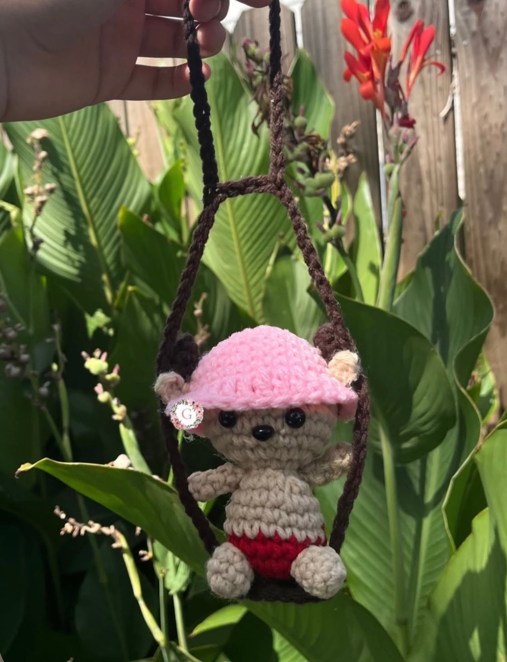 Beige crochet bear wearing a pink hat on a brown swing